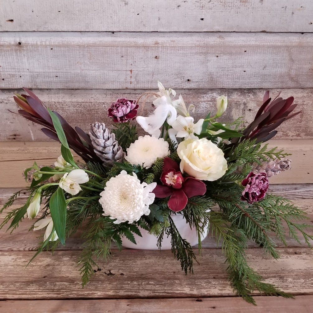 White Roses, Mums, Alstromeria, burgundy Orchids, Luecadendron, carnations with assorted Greenery in a white ceramic container